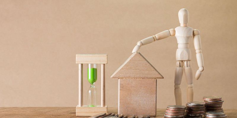 A wooden mannequin figure standing next to a small wooden house model, placing a hand on its roof. Beside the house sits a wooden hourglass with green sand and several scattered and stacked coin piles on a wooden surface, symbolizing mortgage planning, home savings, and timing.