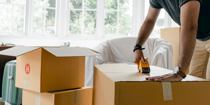 A person using a yellow tape dispenser to seal a cardboard box while packing up a bright, modern living room.