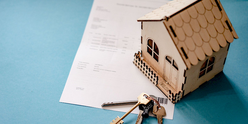 small, detailed miniature house constructed from light-coloured wood sits on the right side of a white document. It features a decorative fence, arched doorway, windows, and a tiled roof pattern. The image symbolizes mortgage approval, property purchasing, renting, or contract signing.